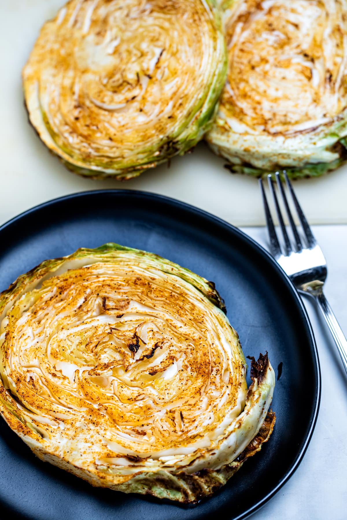 An air fryer cabbage steak on a plate next to a fork with more cabbage steaks on the table.