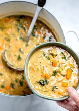 A hand holding a bowl of white bean soup over a pot of soup with a ladle in it.