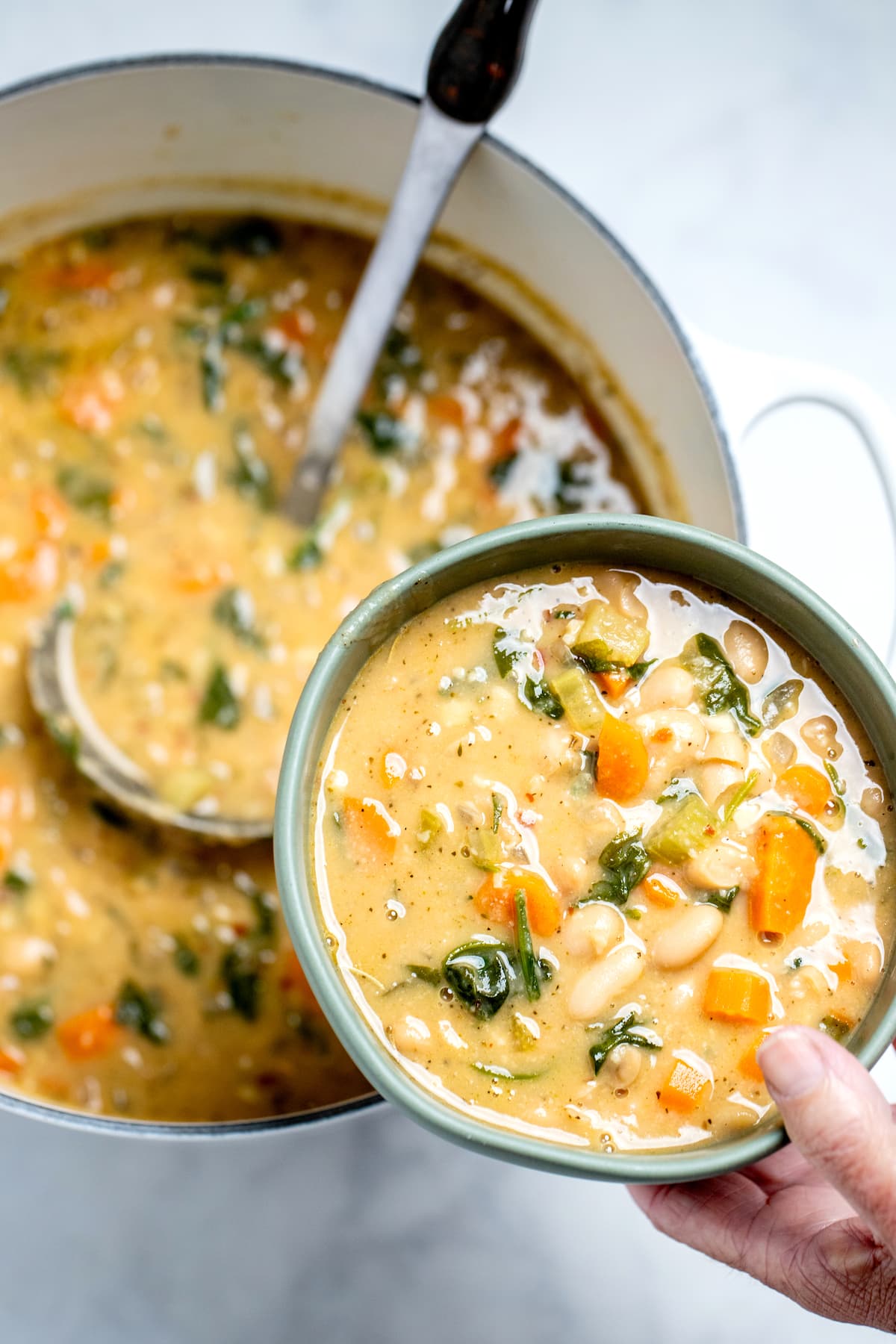 A hand holding a bowl of white bean soup over a pot of soup with a ladle in it.