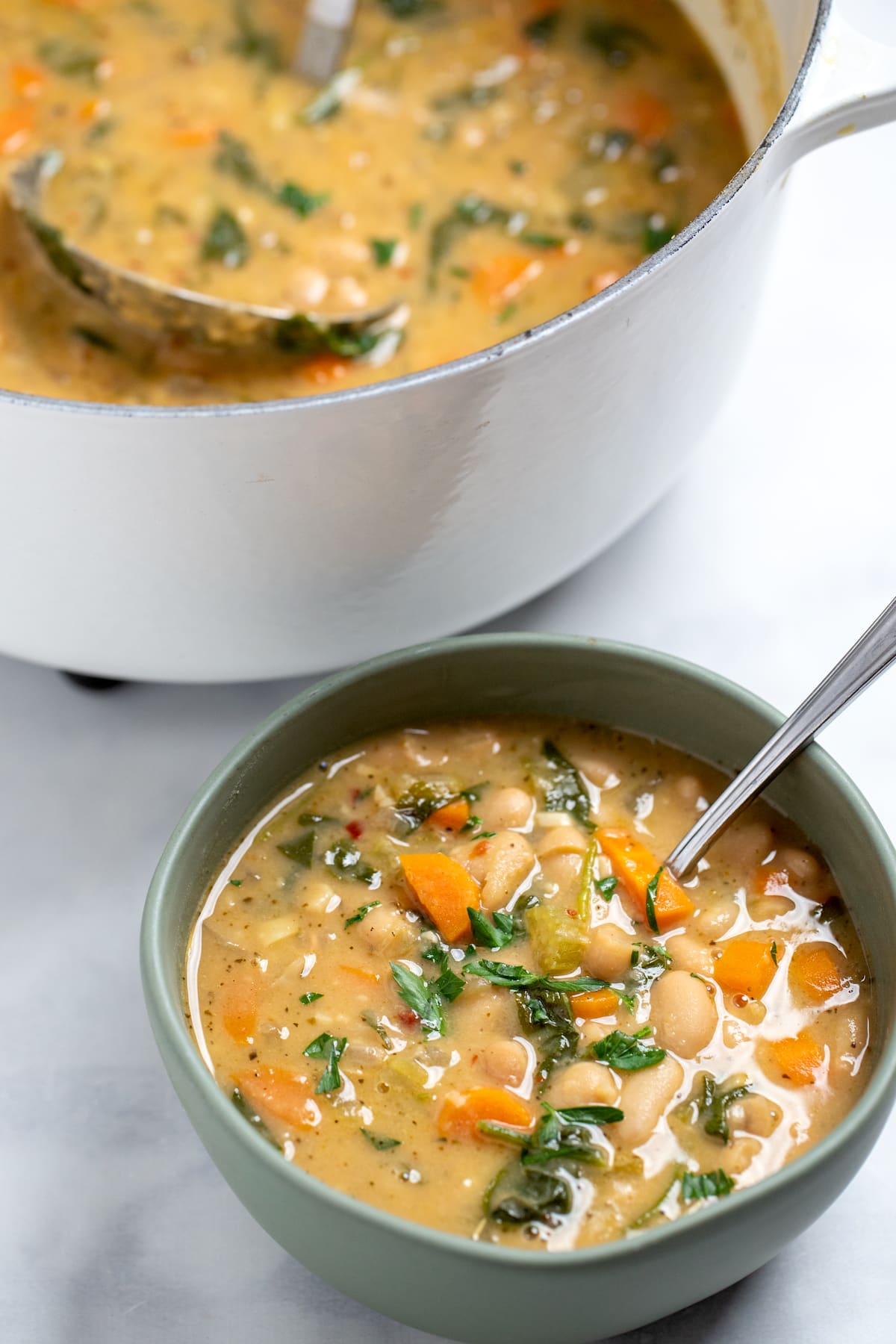 A bowl of tuscan soup with a spoon in front of a dutch oven of soup on a table.