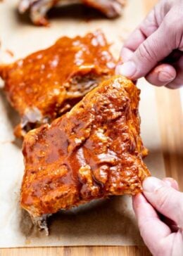 Two hands lifting up a section of pork spare ribs smothered in bbq sauce from a piece of parchment paper on a cutting board, with more sections of ribs in the background.