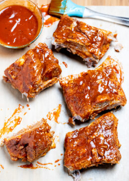 Pork spare ribs cut into sections sitting on parchment paper on top of a cutting board, next to a brush and a bowl of bbq sauce.