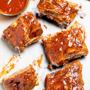 Pork spare ribs cut into sections sitting on parchment paper on top of a cutting board, next to a brush and a bowl of bbq sauce.