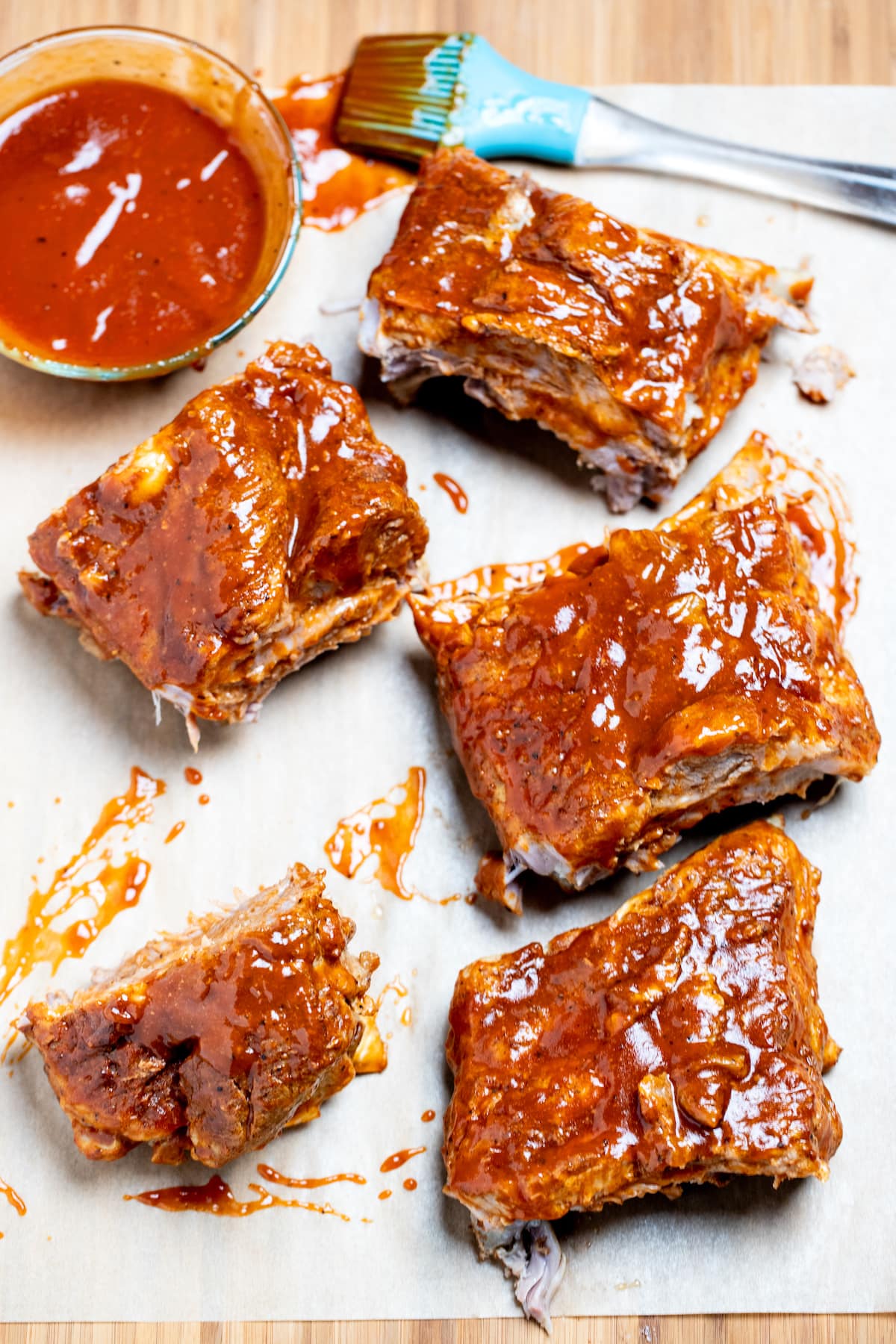 Pork spare ribs cut into sections sitting on parchment paper on top of a cutting board, next to a brush and a bowl of bbq sauce.