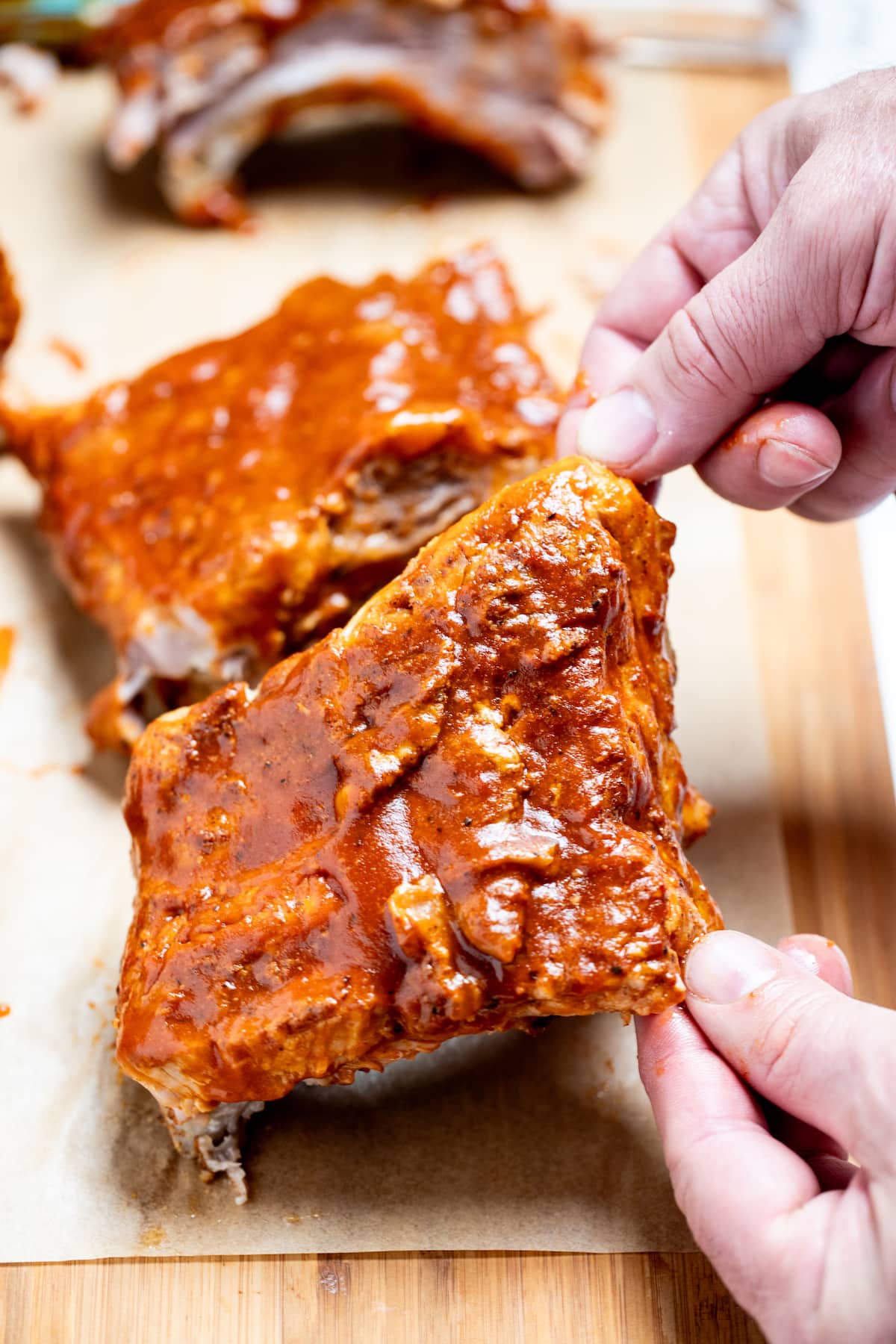Two hands lifting up a section of pork spare ribs smothered in bbq sauce from a piece of parchment paper on a cutting board, with more sections of ribs in the background.