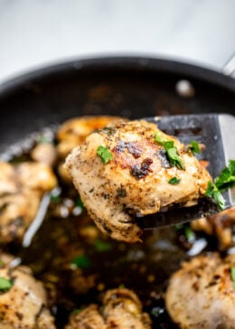 Greek marinated chicken thighs in a skillet topped with fresh parsley, with a spatula lifting a piece of chicken up.