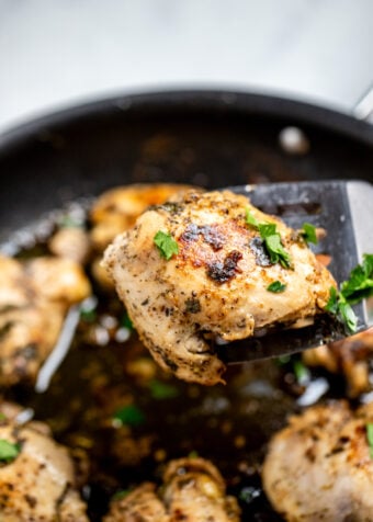 Greek marinated chicken thighs in a skillet topped with fresh parsley, with a spatula lifting a piece of chicken up.