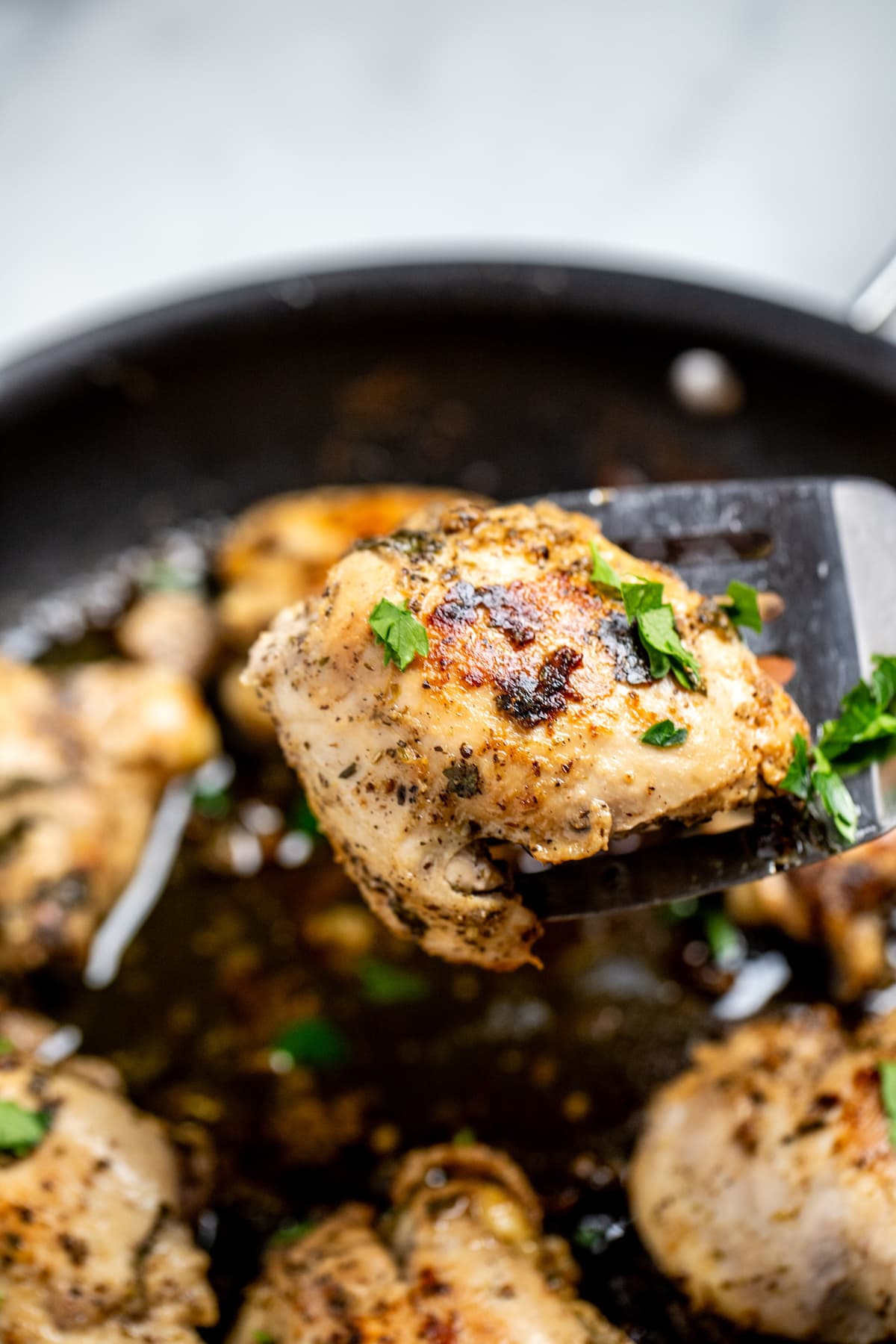 Greek marinated chicken thighs in a skillet topped with fresh parsley, with a spatula lifting a piece of chicken up.