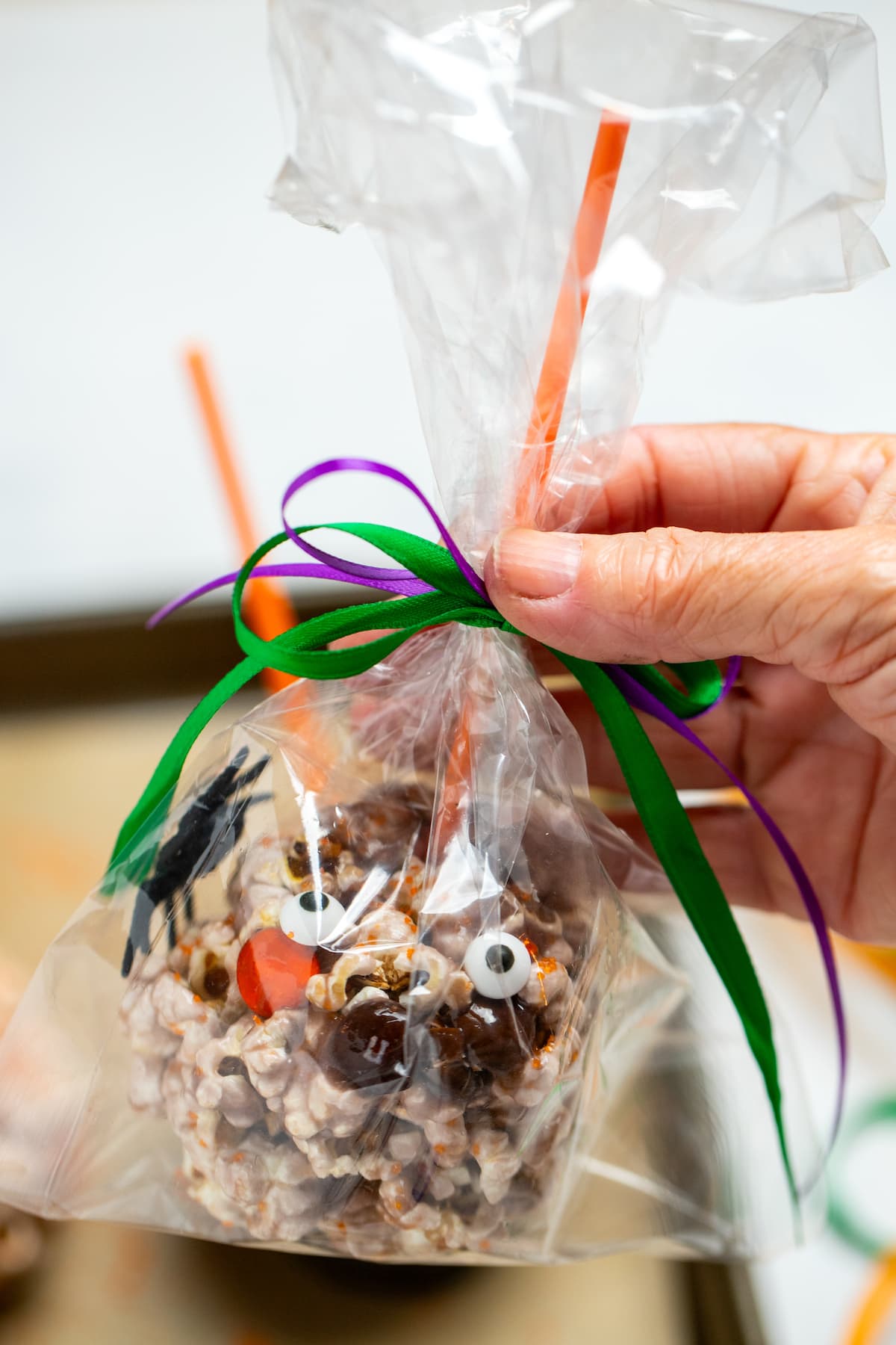 Popcorn ball for halloween with an orange stick on the top, in a plastic gift bag tied with ribbon, being help up by a hand.