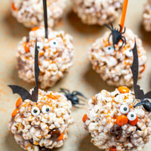 Halloween popcorn balls on a sheet pan surrounded by orange sprinkles and spiders, topped with bat toothpicks.