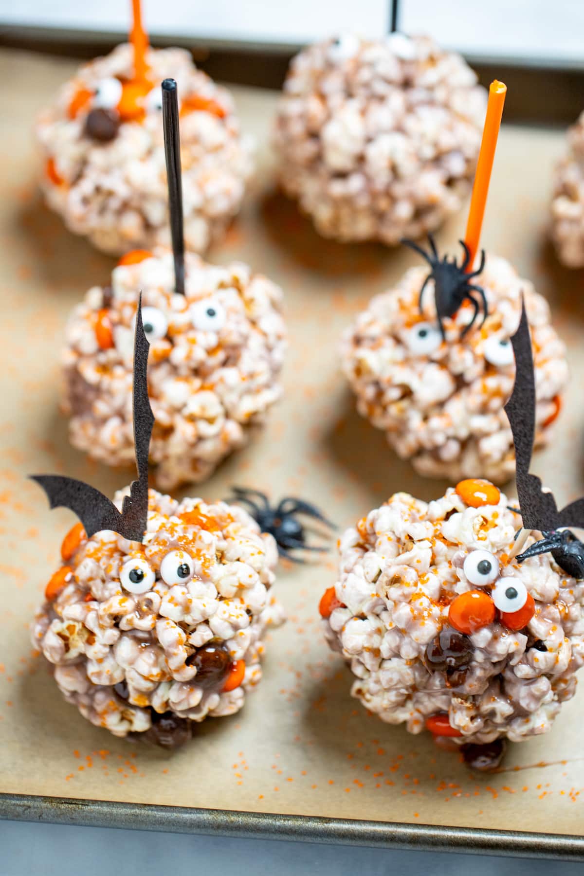 Halloween popcorn balls on a sheet pan surrounded by orange sprinkles and spiders, topped with bat toothpicks.