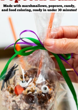Pinterest pin with a popcorn ball for halloween with an orange stick on the top, in a plastic gift bag tied with ribbon, being help up by a hand.