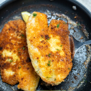 A spatula lifting a piece of parmesan crusted tilapia out of a frying pan with another fillet and lemon wedges in it.