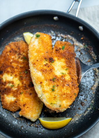 A spatula lifting a piece of parmesan crusted tilapia out of a frying pan with another fillet and lemon wedges in it.