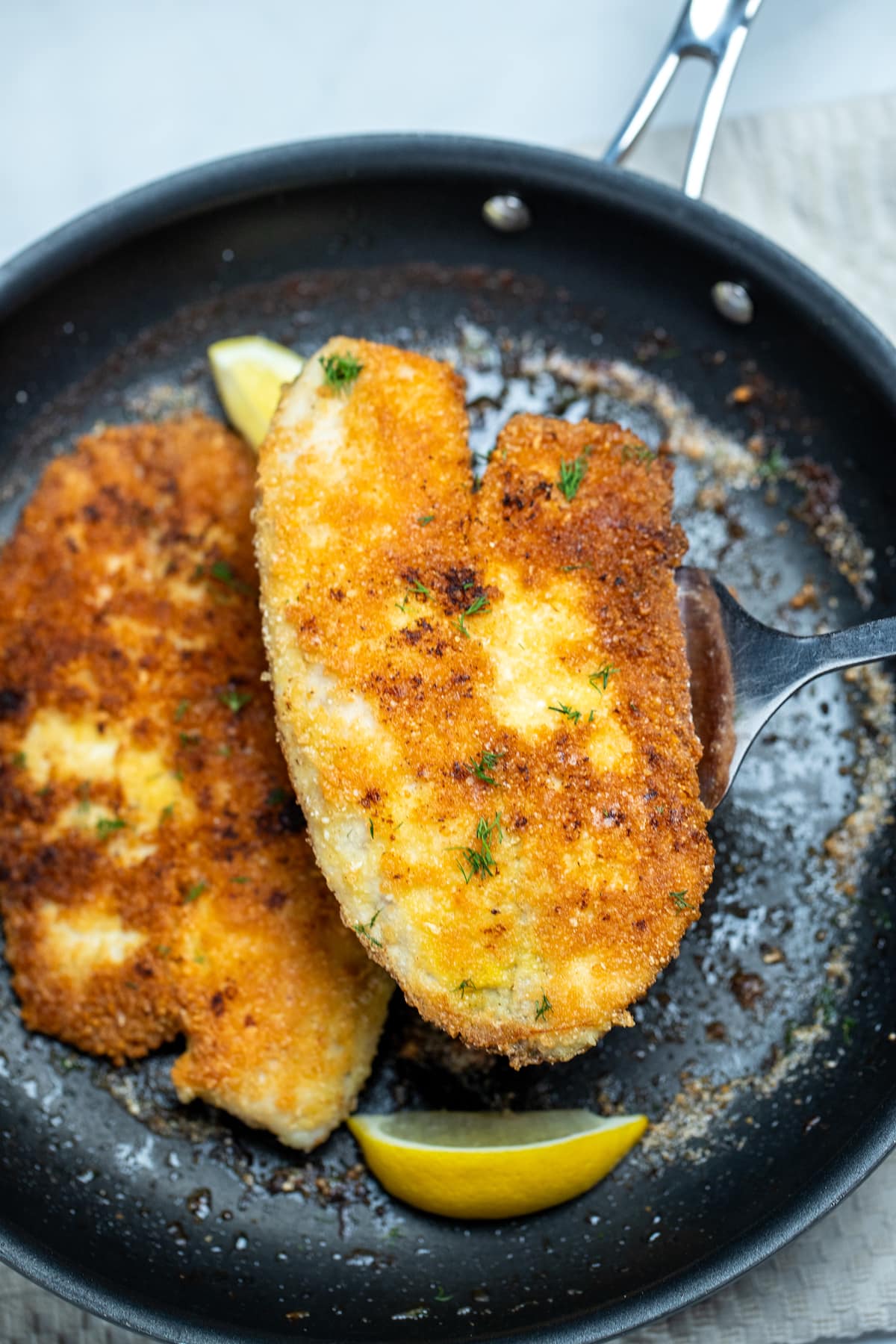 A spatula lifting a piece of parmesan crusted tilapia out of a frying pan with another fillet and lemon wedges in it.