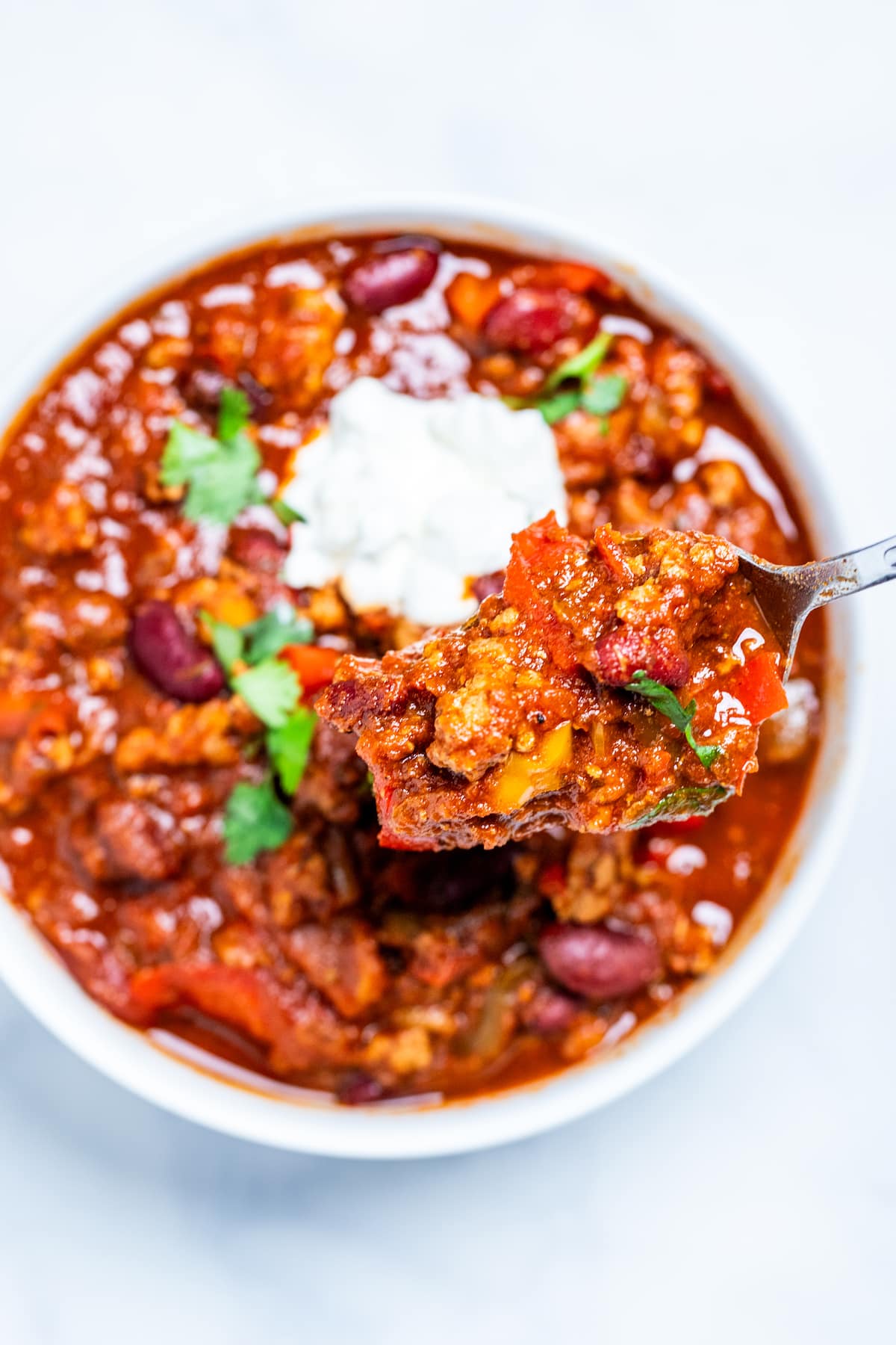 A bowl of crockpot turkey chili topped with sour cream and fresh cilantro, on a table with a spoon scooping some chili out.