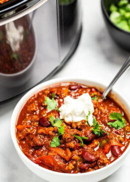 A bowl of slow cooker turkey chili on a table in front of a slow cooker. The chili is topped with fresh cilantro and sour cream.