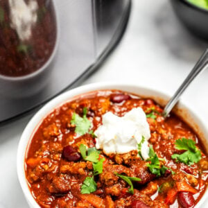 A bowl of slow cooker turkey chili on a table in front of a slow cooker. The chili is topped with fresh cilantro and sour cream.