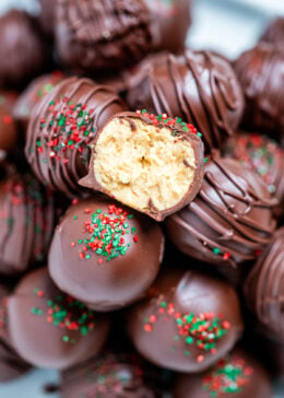 Chocolate covered peanut butter balls piled on a plate, with one cut in half on top, topped with drizzled chocolate and sprinkles on a table.