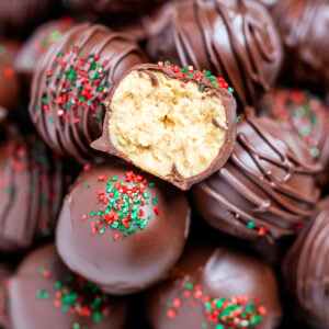 Chocolate covered peanut butter balls piled on a plate, with one cut in half on top, topped with drizzled chocolate and sprinkles on a table.