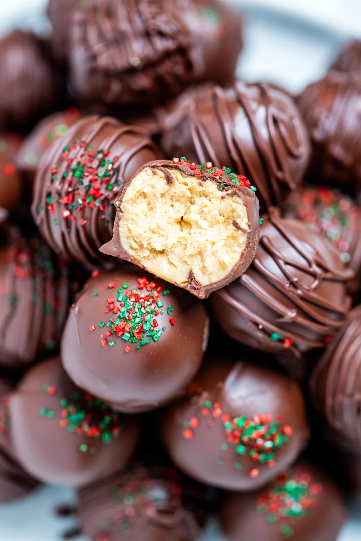 Chocolate covered peanut butter balls piled on a plate, with one cut in half on top, topped with drizzled chocolate and sprinkles on a table.