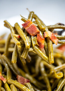 A bowl of crockpot green beans on a table, with a spoon lifting up a scoop of green beans topped with bacon.