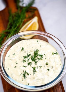 A bowl of homemade tartar sauce on a cutting board, topped with fresh dill, with a lemon wedge in the background.