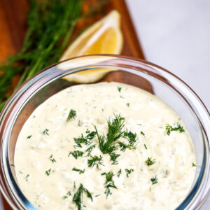 A bowl of homemade tartar sauce on a cutting board, topped with fresh dill, with a lemon wedge in the background.