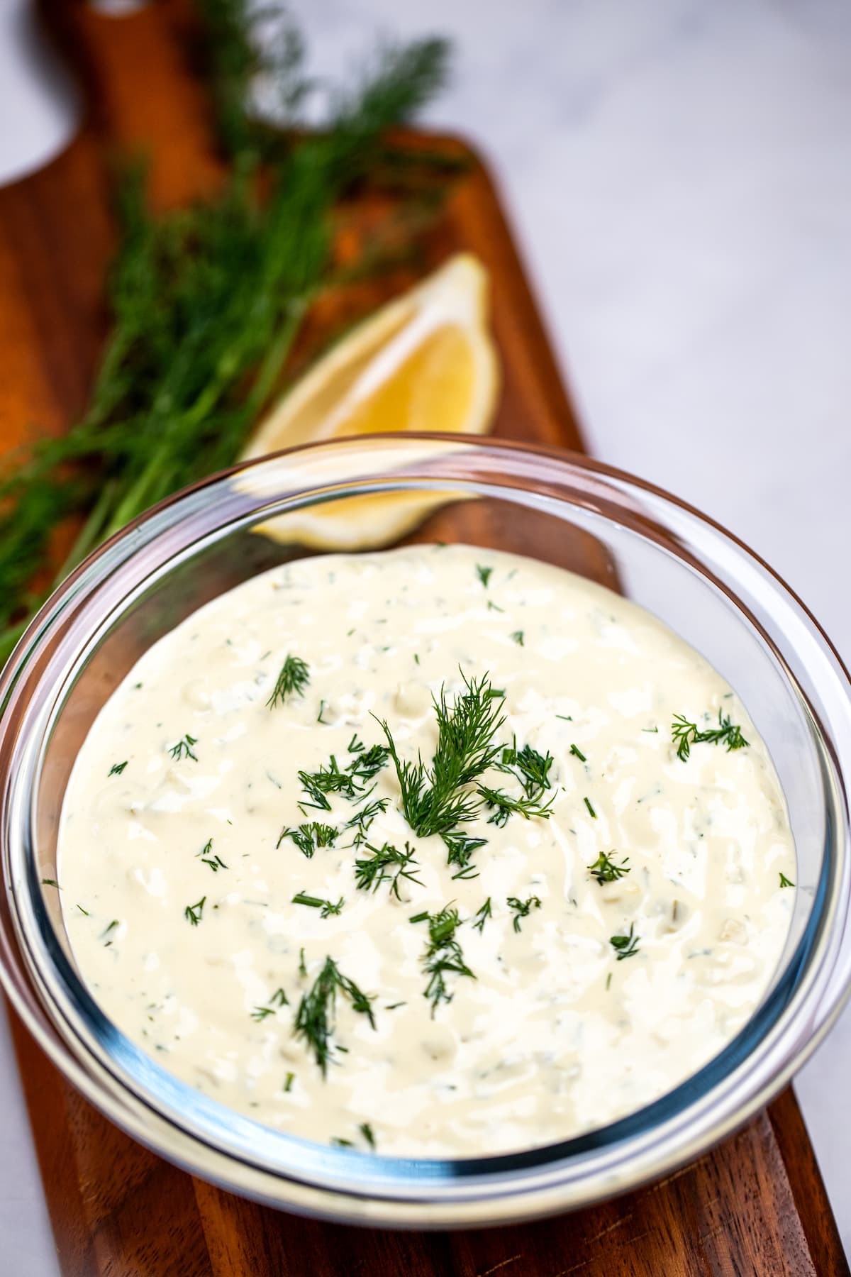 A bowl of homemade tartar sauce on a cutting board, topped with fresh dill, with a lemon wedge in the background.