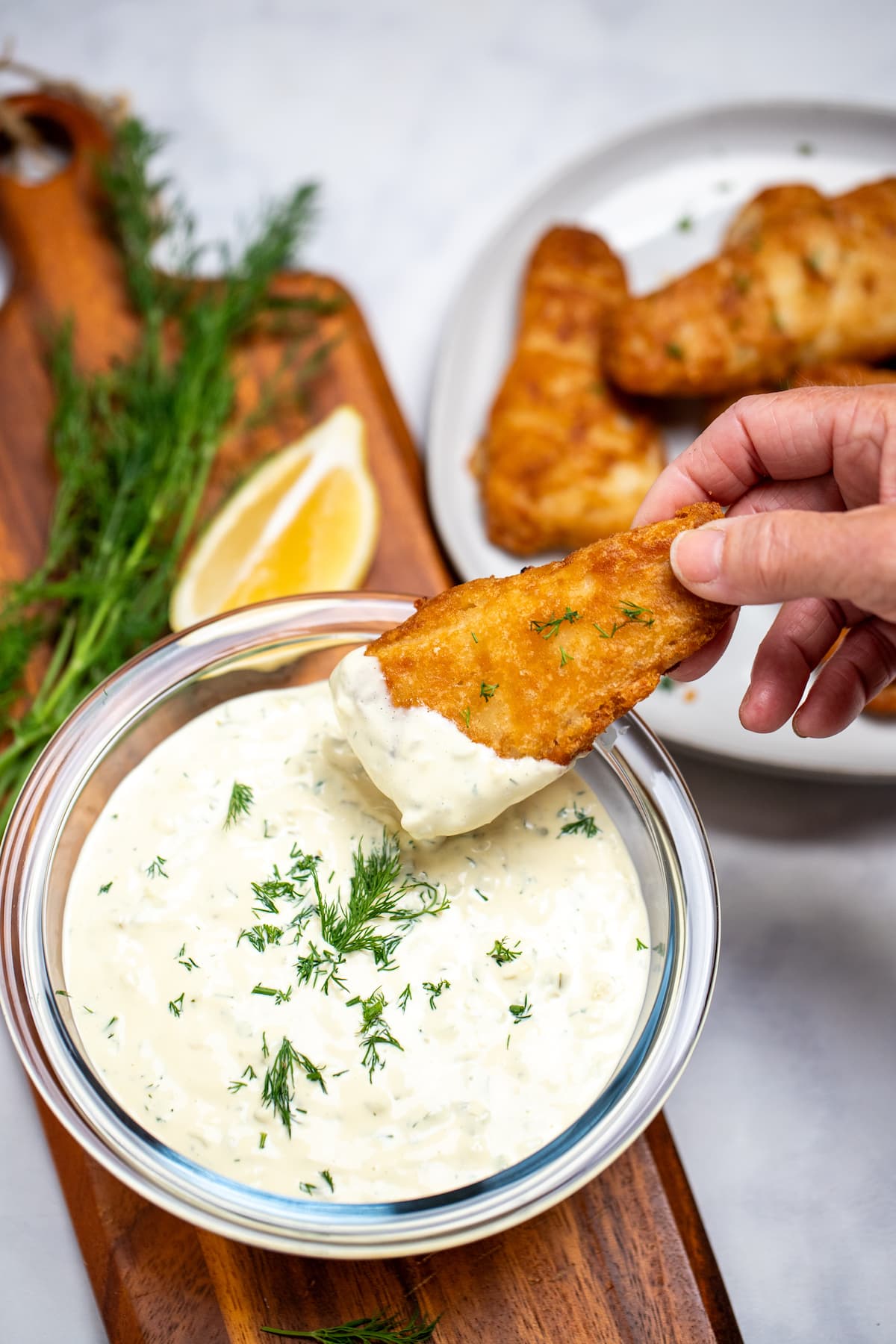 A bowl of homemade tartar sauce on a cutting board, topped with fresh dill and a hand dipping a fish stick into the sauce, with a lemon wedge in the background.