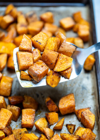Roasted sweet potato cubes on a sheet pan with parchment paper, being lifted up by a spatula.