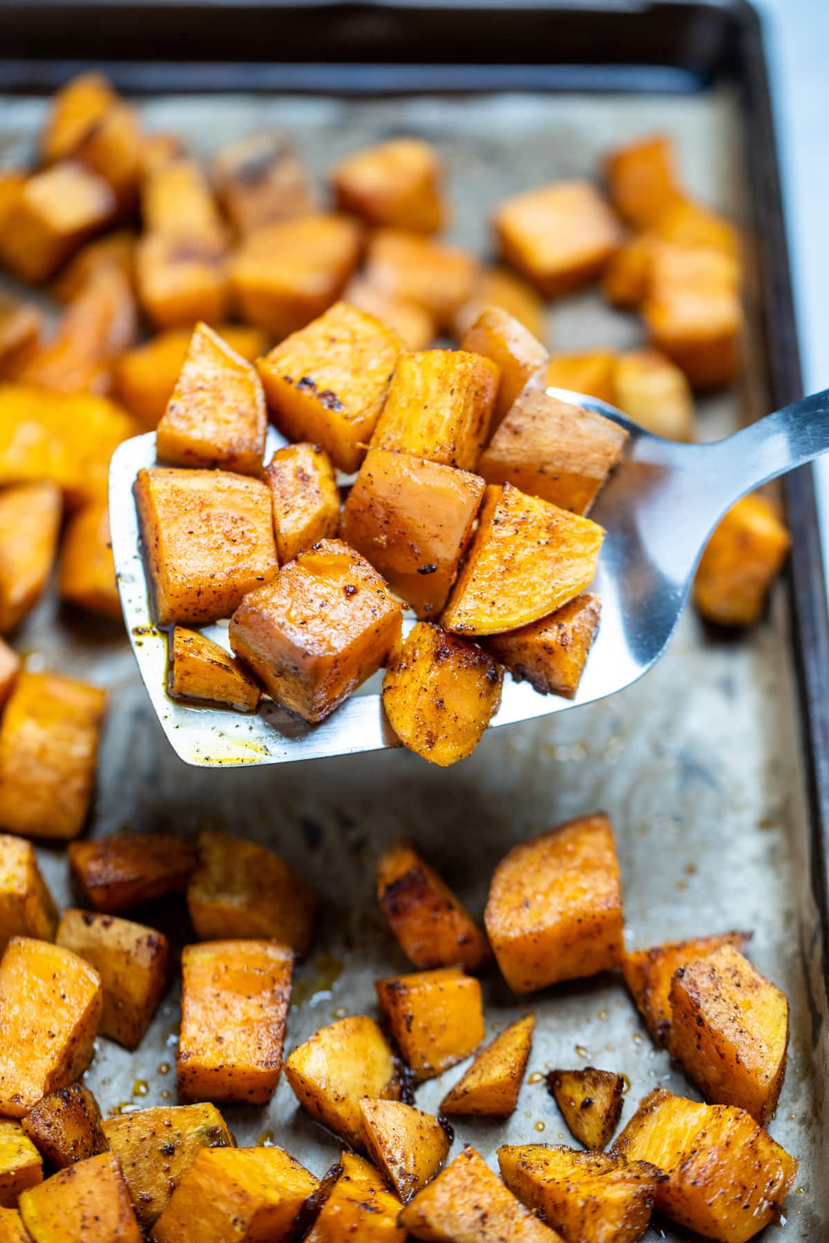 Roasted sweet potato cubes on a sheet pan with parchment paper, being lifted up by a spatula.