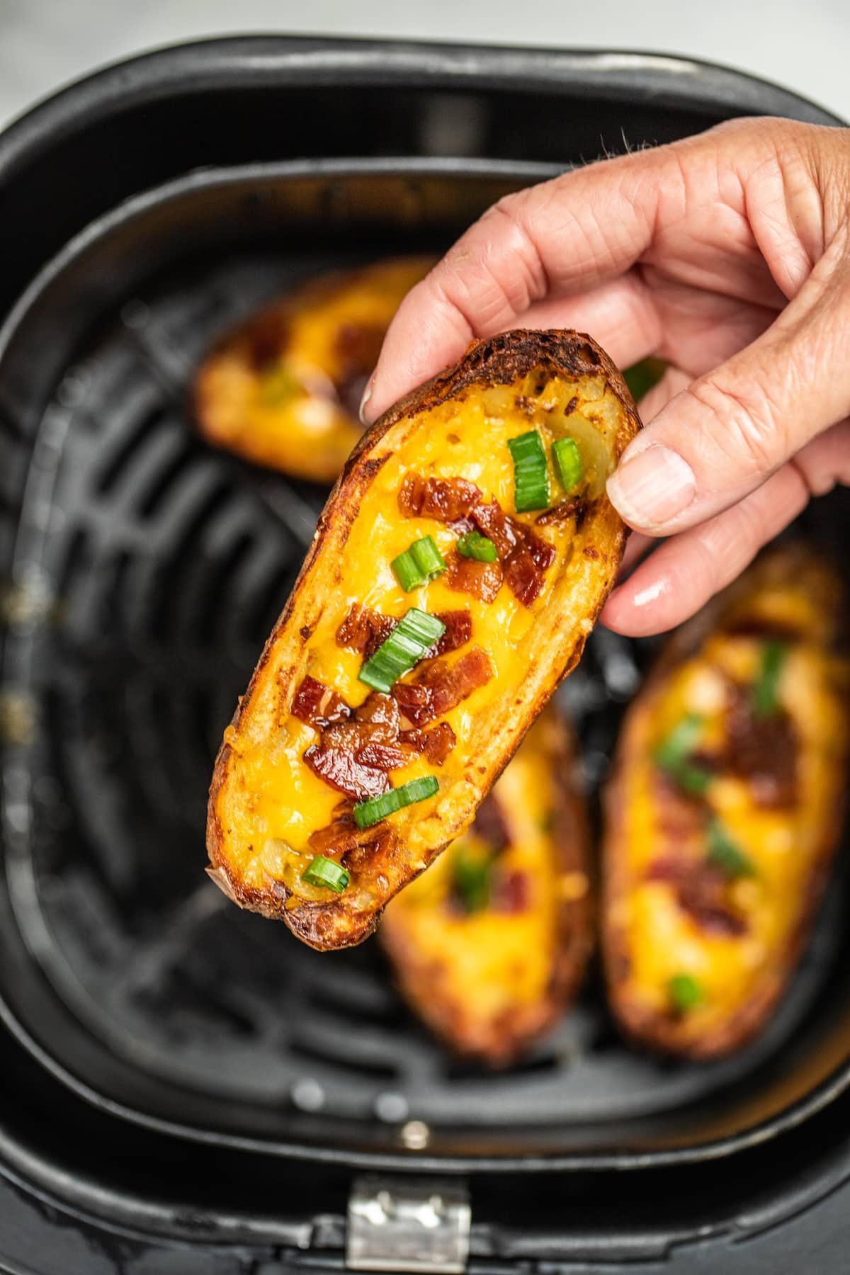 A hand holding a potato skin above an air fryer basket of cooked potato skins.