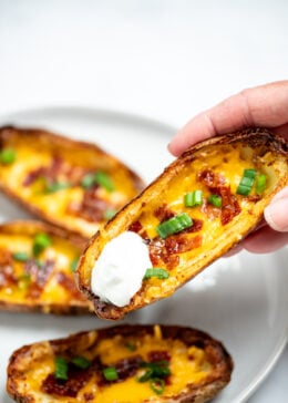 A hand holding a potato skin with sour cream above a plate of more potato skins.