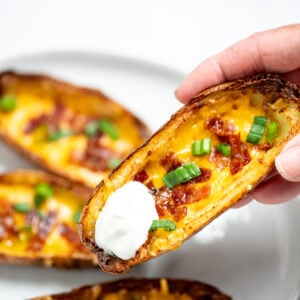 A hand holding a potato skin with sour cream above a plate of more potato skins.