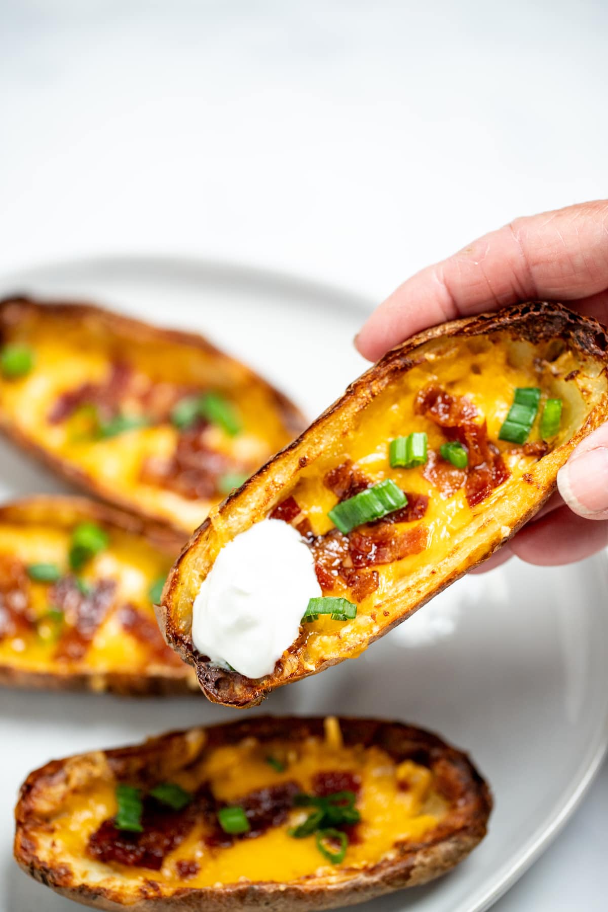 A hand holding a potato skin with sour cream above a plate of more potato skins.