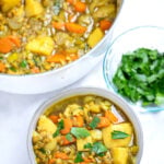 A bowl of lentil soup with carrots, potatoes, lentils, and leeks topped with chopped parsley on a table in front of a dutch oven of soup and a small bowl of fresh chopped parsley.