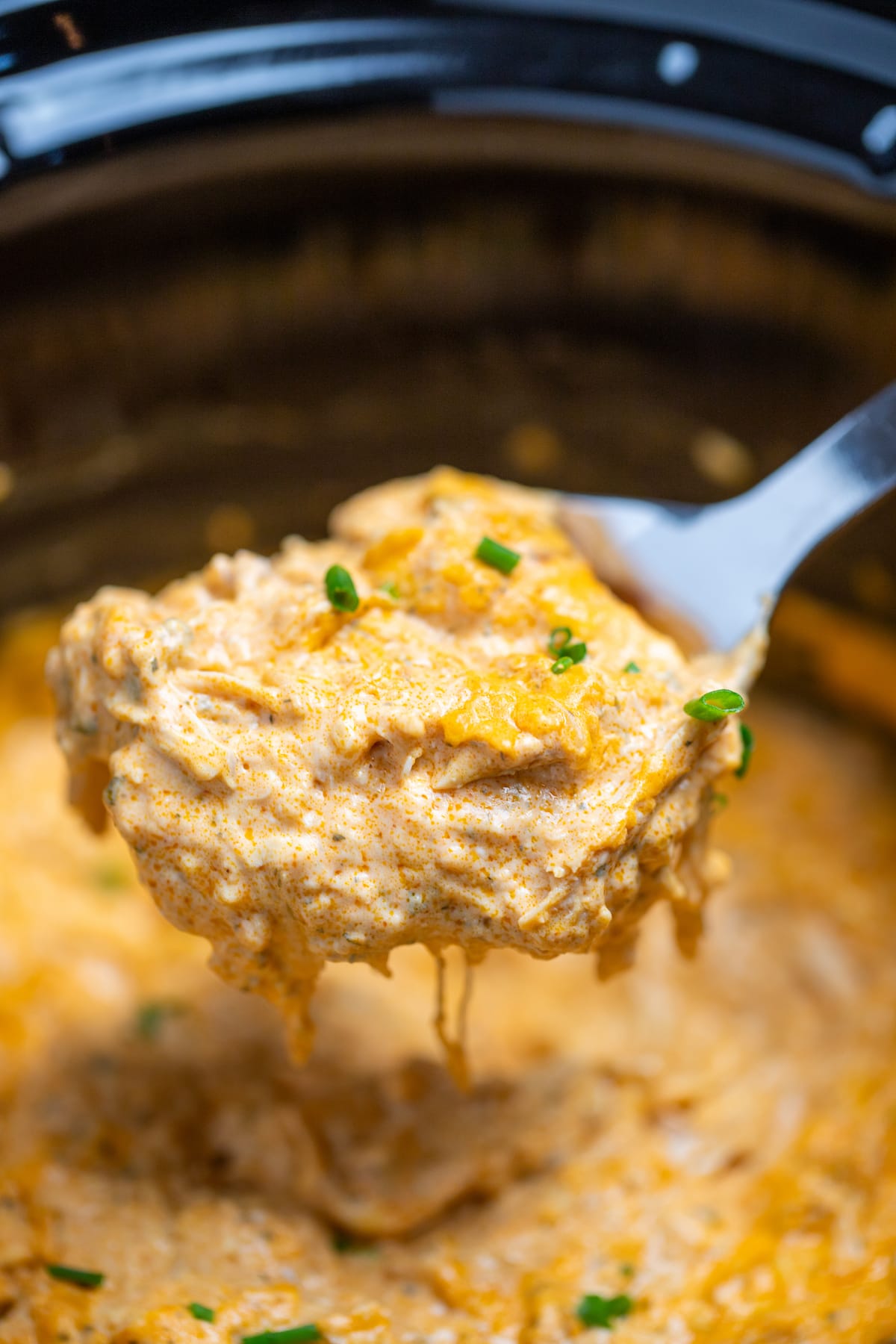 A spoon lifting buffalo chicken dip topped with chives out of the slow cooker.