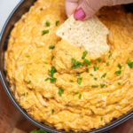 A bowl of slow cooker buffalo chicken dip topped with chives on a wooden cutting board next to celery and tortilla chips, with a hand dipping a chip into the dip.