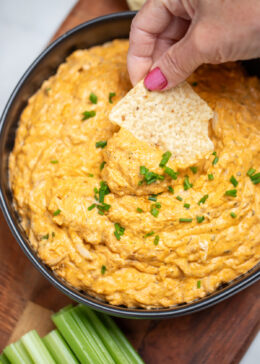 A bowl of slow cooker buffalo chicken dip topped with chives on a wooden cutting board next to celery and tortilla chips, with a hand dipping a chip into the dip.