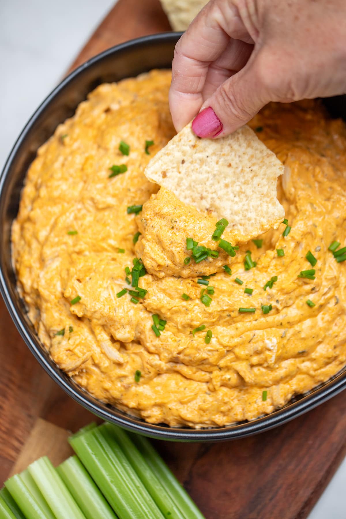 A bowl of slow cooker buffalo chicken dip topped with chives on a wooden cutting board next to celery and tortilla chips, with a hand dipping a chip into the dip.