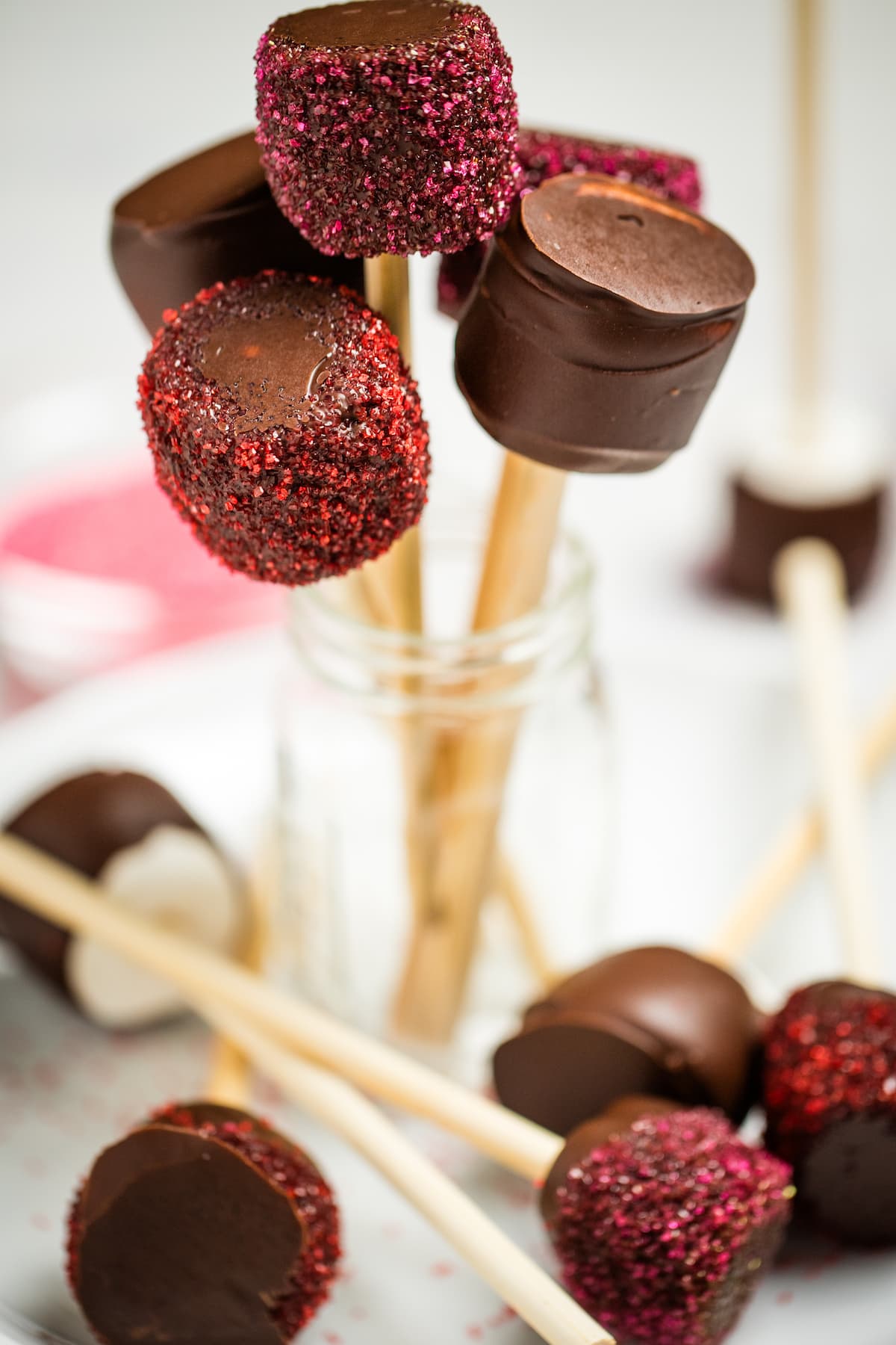 A jar with chocolate dipped marshmallows rolled in sprinkles, with the marshmallows on top on lollipop sticks, behind more marshmallows laying on a plate.