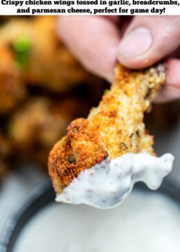 Pinterest pin with a hand holding a garlic parmesan chicken wing after dipping it in ranch, with more wings in the background.