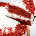 A double-layer gluten free red velvet cake on a wooden cake plate with a piece of cake being lifted up.