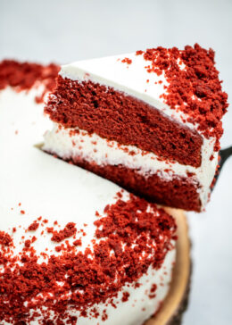 A double-layer gluten free red velvet cake on a wooden cake plate with a piece of cake being lifted up.