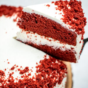 A double-layer gluten free red velvet cake on a wooden cake plate with a piece of cake being lifted up.