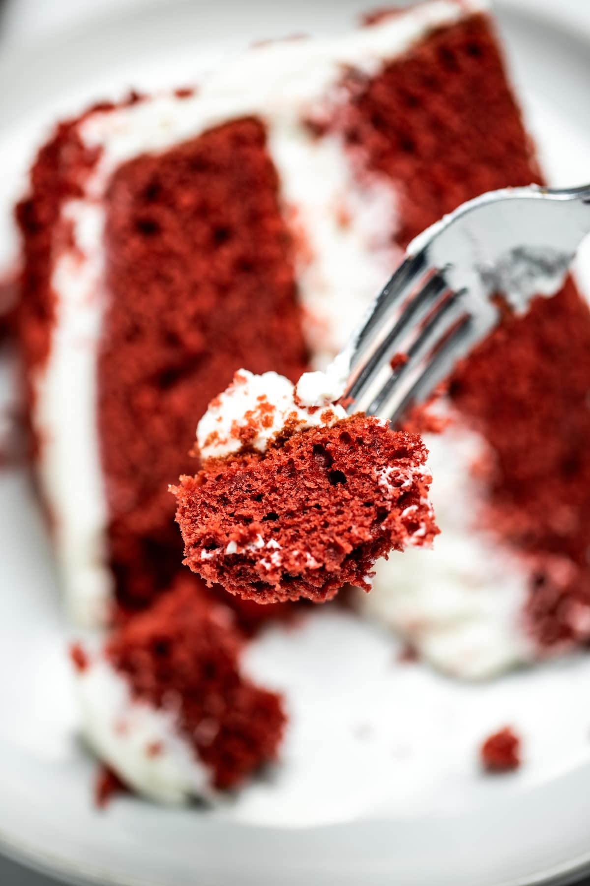 A closeup of a fork with a piece of red velvet cake, with a piece of cake on a plate in the background.