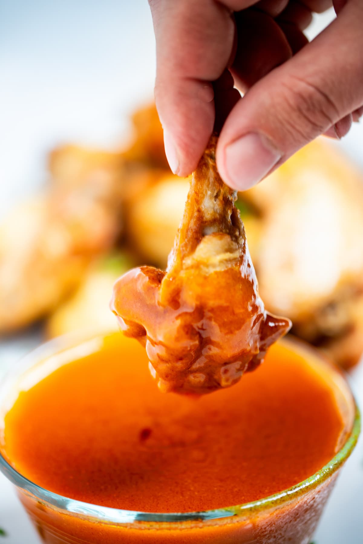 A hand holding a chicken wing after dipping it into a bowl of homemade buffalo wing sauce, with a plate of wings in the background.