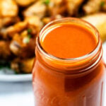 A mason jar full of homemade buffalo wing sauce in front of a plate of chicken wings.