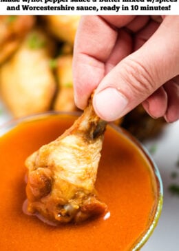 A hand holding a chicken wing while dipping it into a bowl of homemade buffalo wing sauce, with a plate of wings in the background.